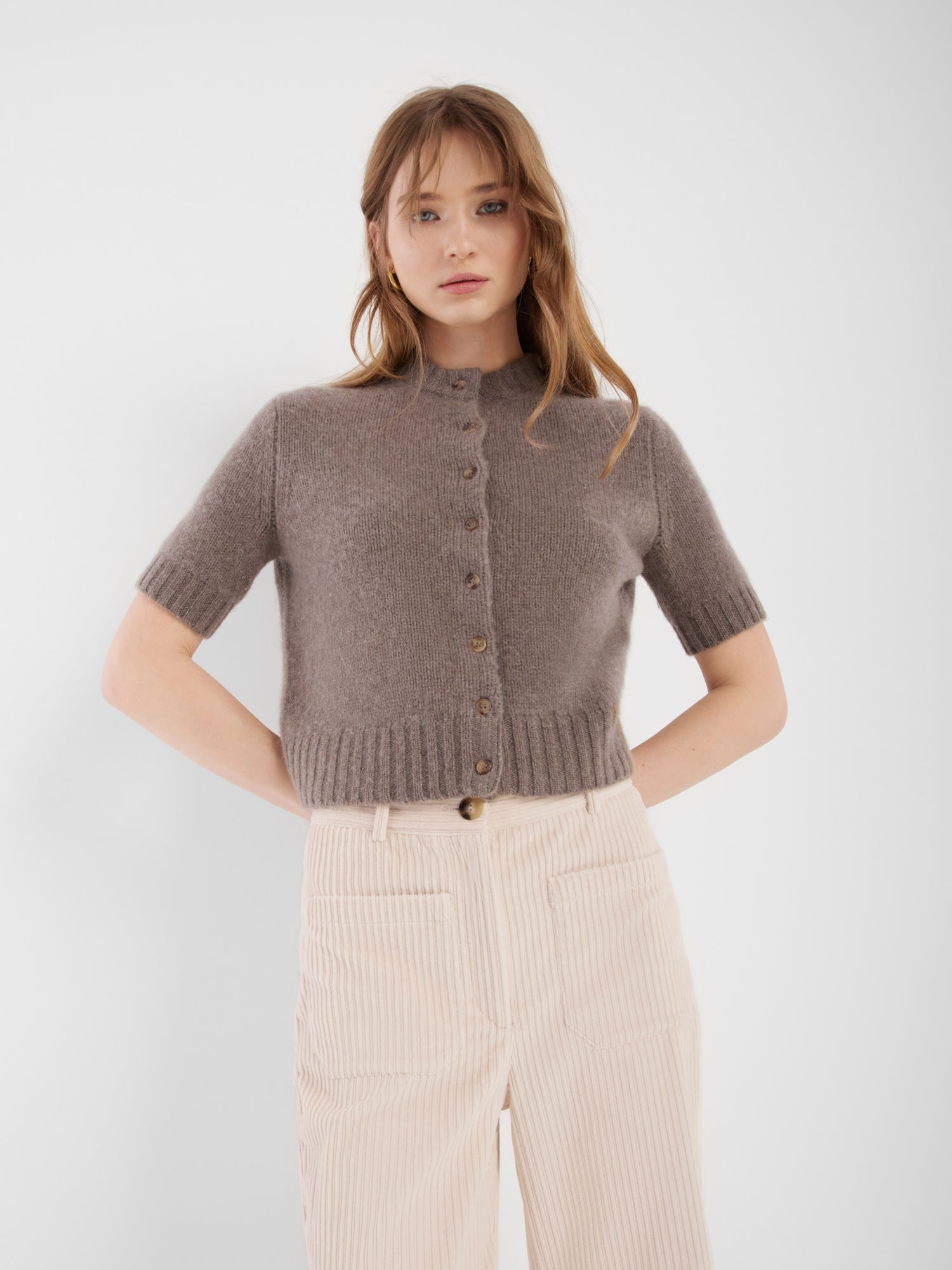 Eine Frau mit hellbraunem Haar trägt einen kurzärmeligen Azteken-Pullover in Taupebraun und eine hochtaillierte, hellbeige Hose. Sie steht vor einem schlichten weißen Hintergrund und hat die Hände in die Hüften gestemmt.