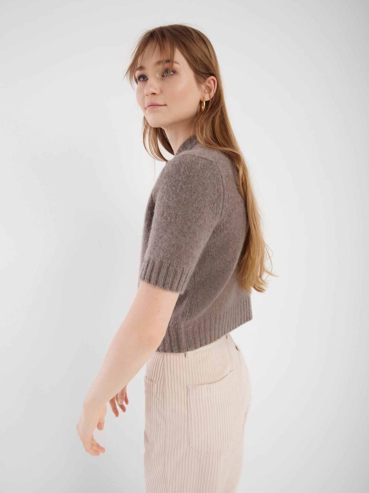 Eine Frau mit langem, hellbraunem Haar trägt einen kurzärmeligen Azteken-Pullover und eine hochtaillierte, hellbeige Hose. Sie steht seitlich vor einem schlichten weißen Hintergrund und schaut leicht über ihre Schulter.