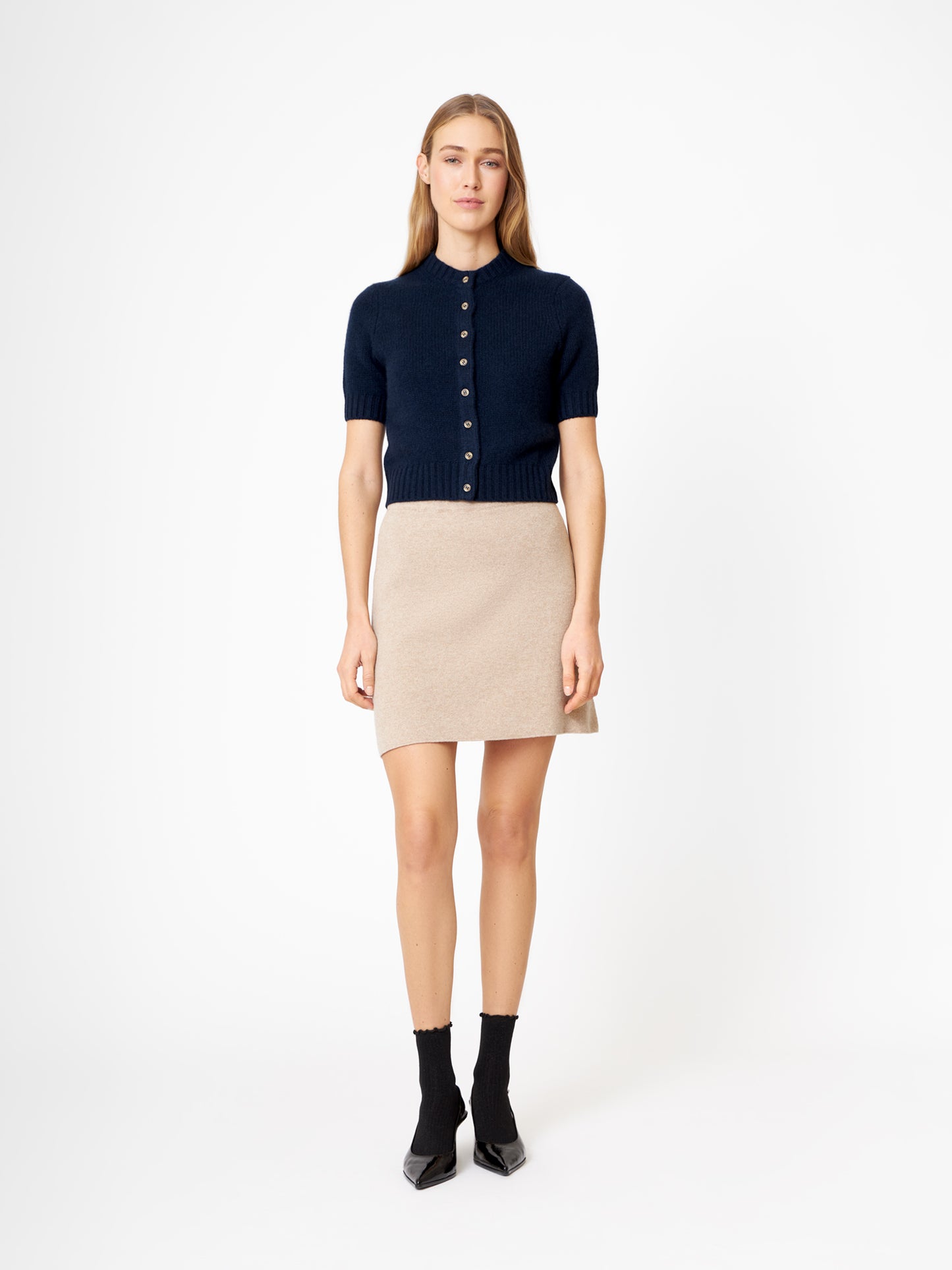Eine Frau steht mit dem Gesicht nach vorne und trägt einen dunkelblauen Kaschmirpullover mit kurzen Ärmeln, einen beigen Minirock, schwarze Socken und schwarze spitze Schuhe vor einem schlichten weißen Hintergrund.