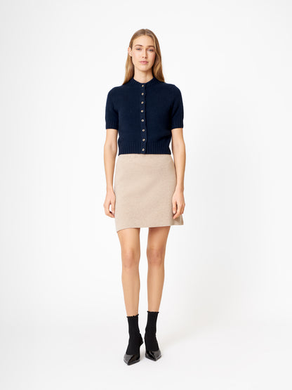 Eine Frau steht mit dem Gesicht nach vorne und trägt einen dunkelblauen Kaschmirpullover mit kurzen Ärmeln, einen beigen Minirock, schwarze Socken und schwarze spitze Schuhe vor einem schlichten weißen Hintergrund.