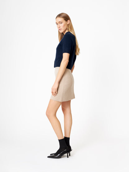 Eine Frau mit langen blonden Haaren steht im Profil vor einem weißen Hintergrund und trägt einen dunkelblauen Kaschmir-Pullover mit kurzen Ärmeln, einen beigen Minirock, schwarze Socken und schwarze Schuhe mit Absatz.