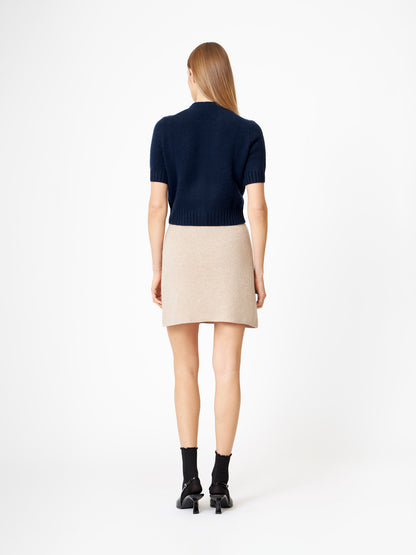 Eine Frau mit glattem blondem Haar steht mit dem Gesicht zur Wand und trägt einen dunkelblauen Kaschmir-Pullover mit kurzen Ärmeln, einen beigen Minirock, schwarze Socken und schwarze hochhackige Sandalen vor einem schlichten weißen Hintergrund.