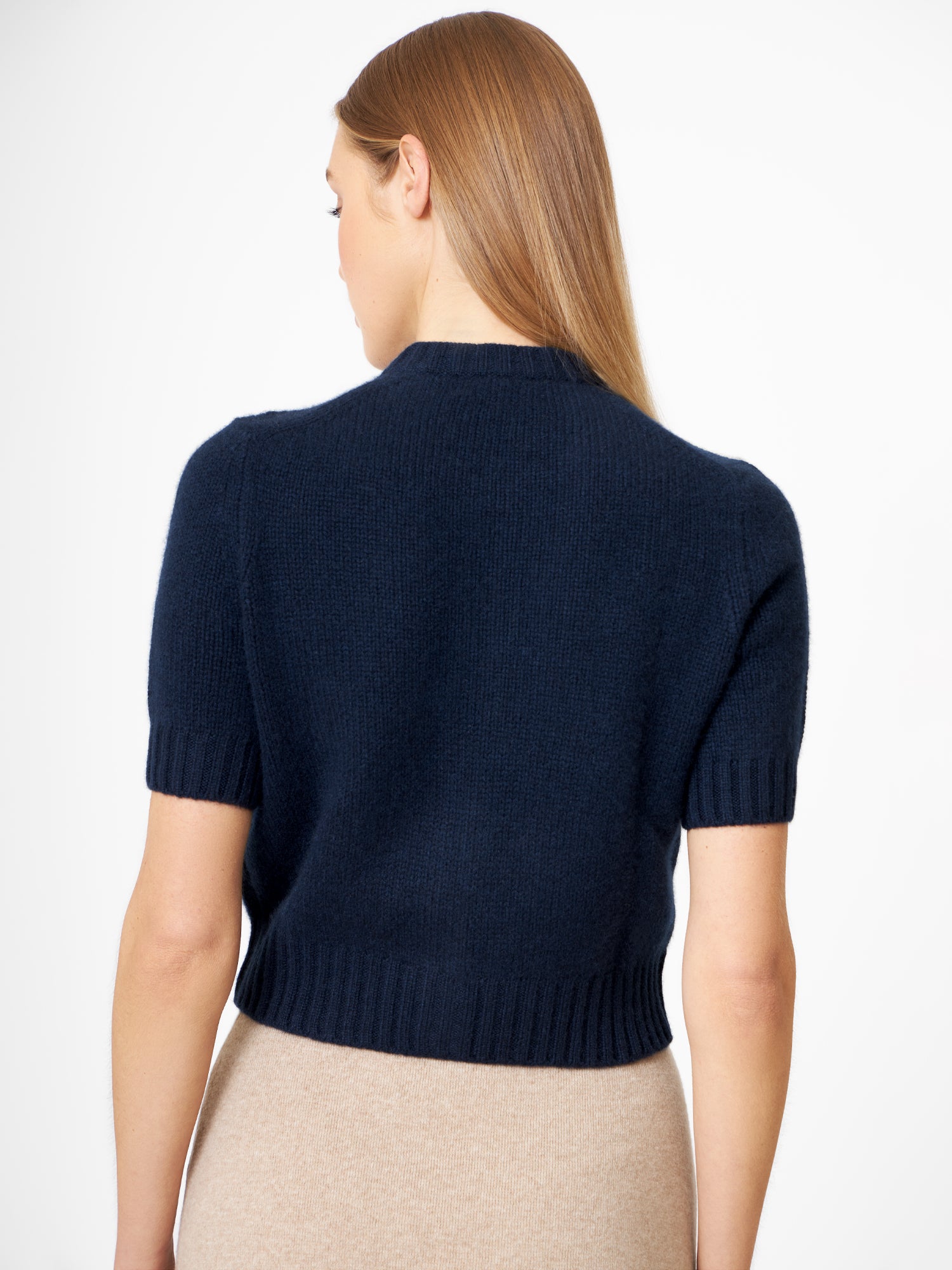 Eine Frau mit glattem, hellbraunem Haar trägt einen dunkelblauen Kaschmirpullover mit kurzen Ärmeln und einen beigen Rock. Sie blickt von der Kamera weg, wodurch die Rückseite des Pullovers und ihr Haar hervorgehoben werden.