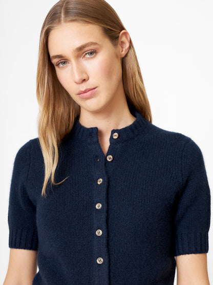 Eine Frau mit langen, hellbraunen Haaren trägt einen Kaschmir-Pullover mit kurzen Ärmeln in Dunkelblau, der als Strickjacke gestylt ist. Sie steht vor einem weißen Hintergrund und blickt mit neutralem Blick leicht zur Seite.