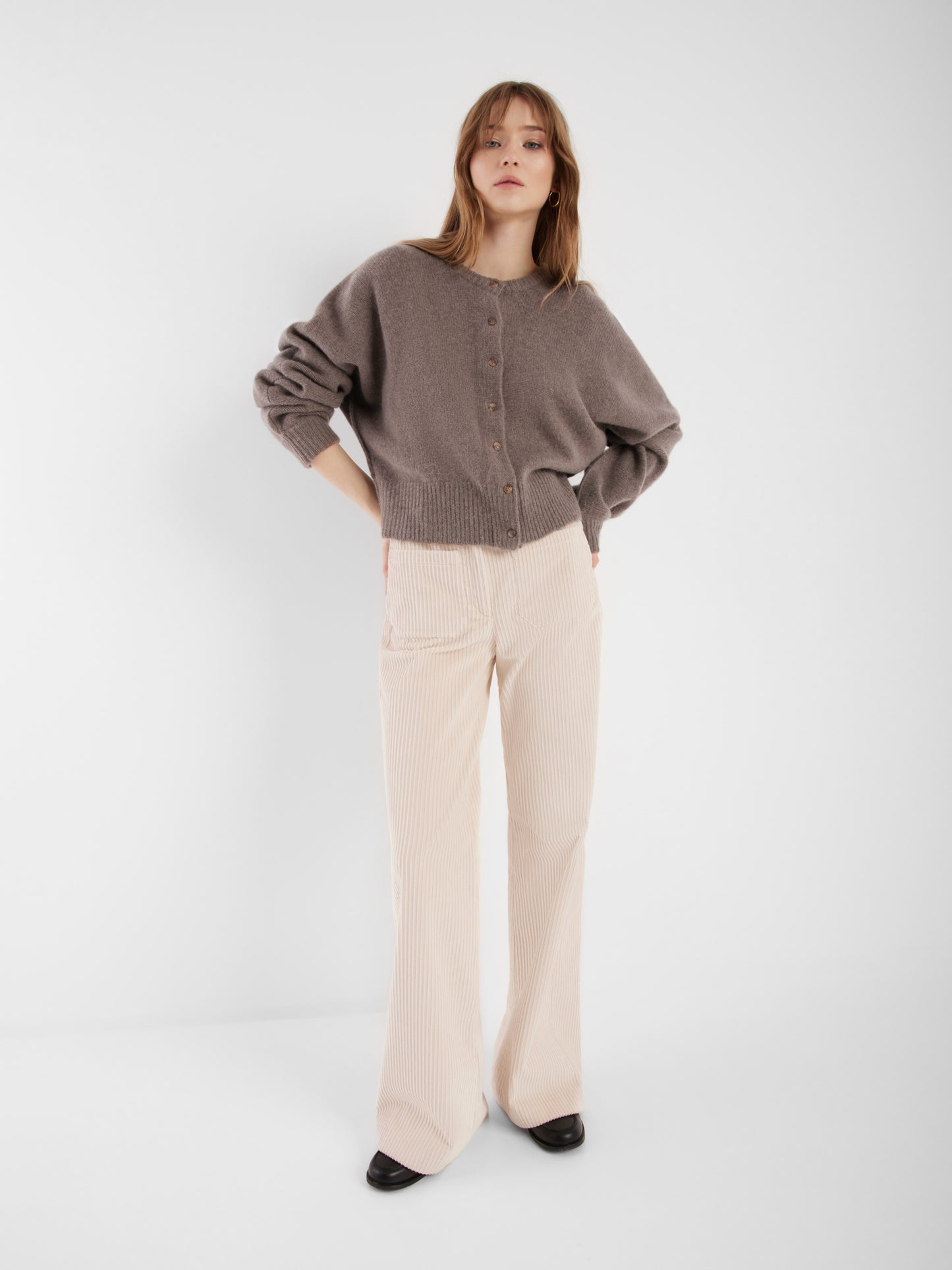 Eine Frau posiert selbstbewusst vor einem schlichten weißen Hintergrund. Sie trägt die cremefarbene Jacke Cashmere Azteco und eine cremefarbene Hose mit weitem Bein und schwarzen Schuhen, die Hände in die Hüften gestützt.