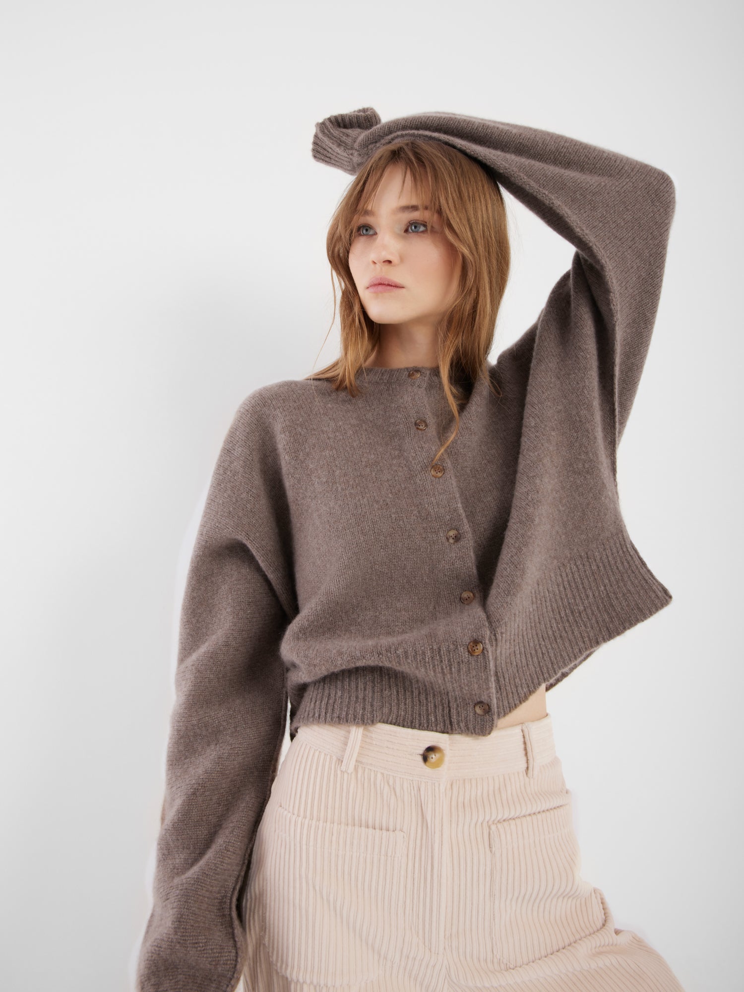 Eine Frau mit hellbraunem Haar posiert mit erhobenem Arm. Sie trägt die locker sitzende, kurz geschnittene Jacke Cashmere Azteco und eine cremefarbene Cordhose mit hoher Taille vor einem schlichten weißen Hintergrund.