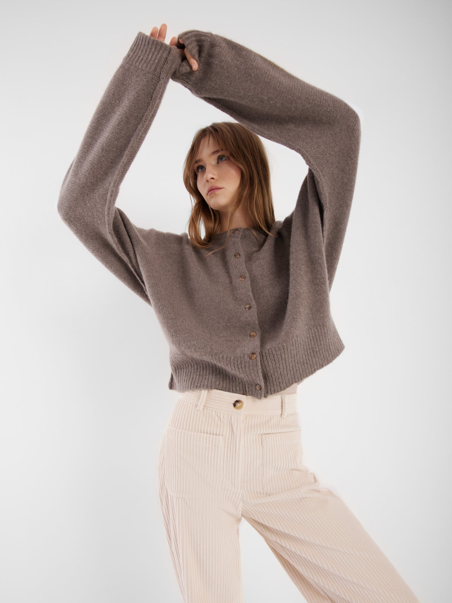 Eine Frau, die die lockere braune Jacke Cashmere Azteco und eine hochtaillierte cremefarbene Cordhose trägt, posiert mit erhobenen und über dem Kopf verschränkten Armen vor einem schlichten weißen Hintergrund.