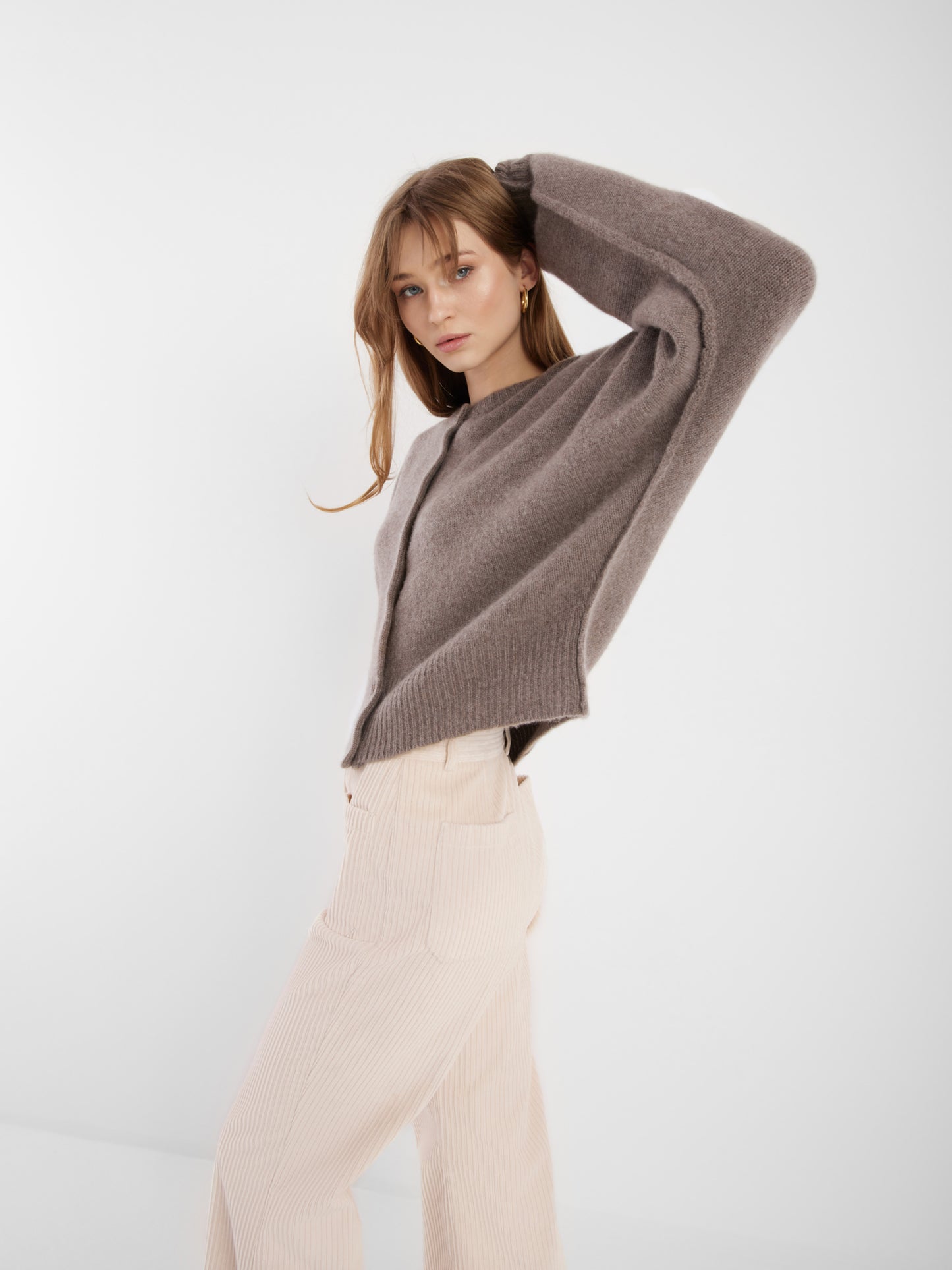 Eine Frau in einer cremefarbenen Jacke Cashmere Azteco und einer cremefarbenen Hose posiert mit einem erhobenen Arm vor einem schlichten weißen Hintergrund.