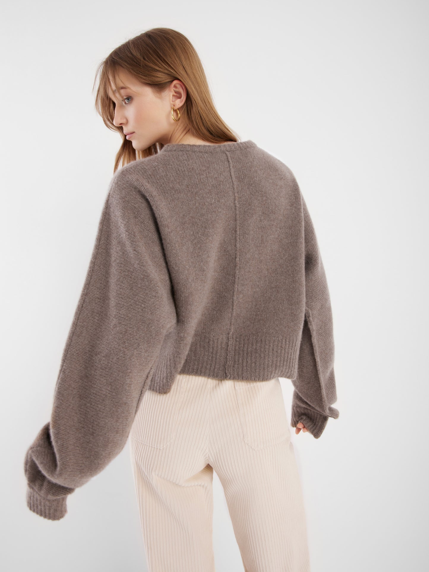 Eine Frau mit hellbraunem Haar trägt eine lockere, taupefarbene Kaschmir-Aztekenjacke mit überschnittenen Schultern und langen Ärmeln, kombiniert mit einer hochtaillierten cremefarbenen Cordhose. Sie ist leicht von der Kamera weggedreht.