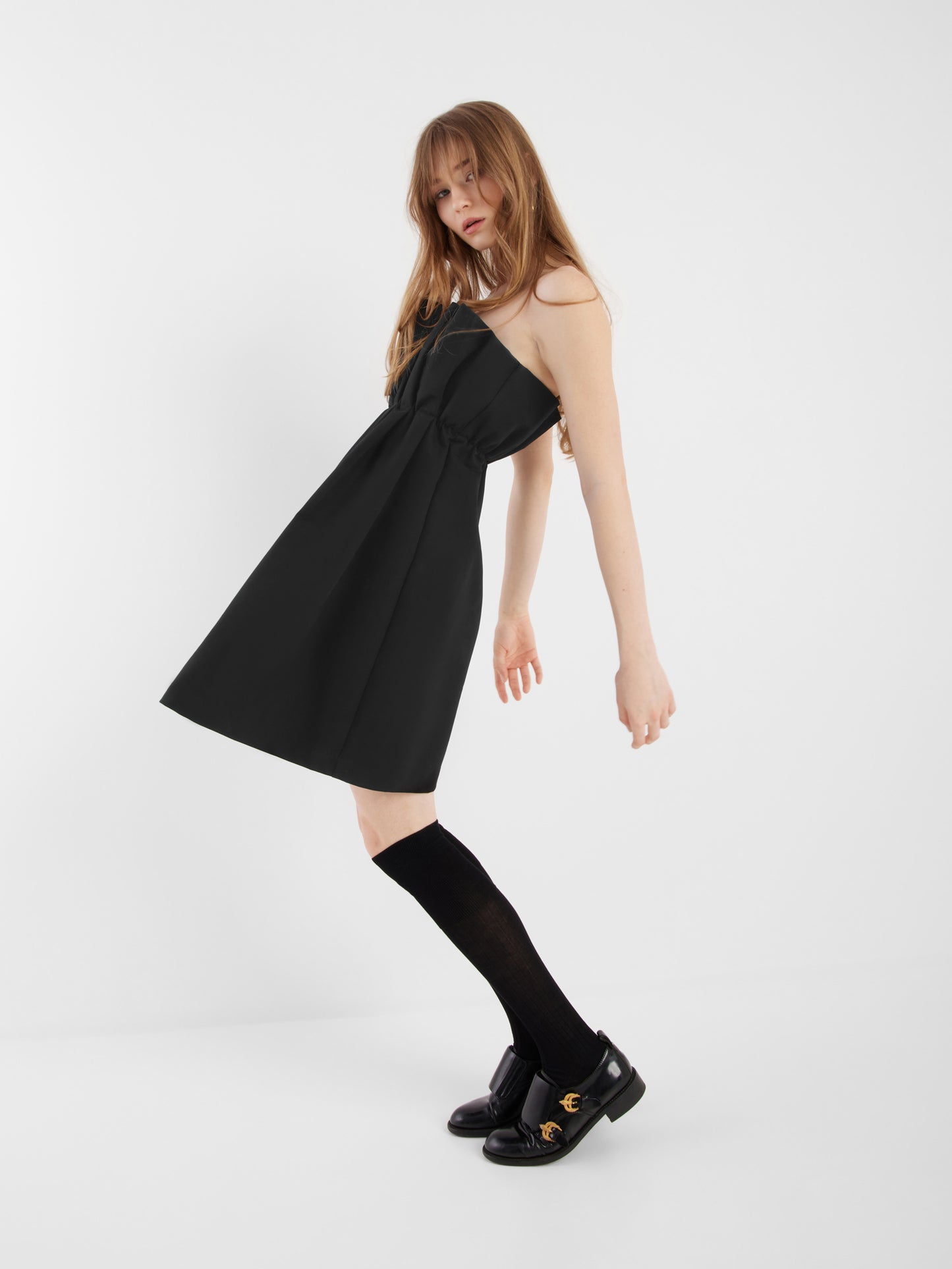 Eine Person mit langen Haaren lehnt sich vor einem einfarbig weißen Hintergrund leicht zur Seite. Sie trägt die Kleiderfalte schwarz, gepaart mit schwarzen Kniestrümpfen und schwarzen Halbschuhen.