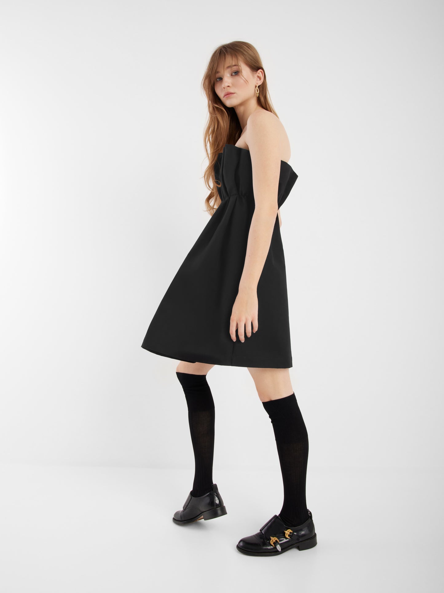 Eine Frau mit langem, hellbraunem Haar trägt das schwarze Kleid Pleat, schwarze Kniestrümpfe und schwarze Slipper mit goldenen Details. Sie steht in einem weißen, minimalistischen Raum und blickt in die Kamera.