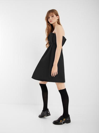 Eine Frau mit langem, hellbraunem Haar trägt das schwarze Kleid Pleat, schwarze Kniestrümpfe und schwarze Slipper mit goldenen Details. Sie steht in einem weißen, minimalistischen Raum und blickt in die Kamera.