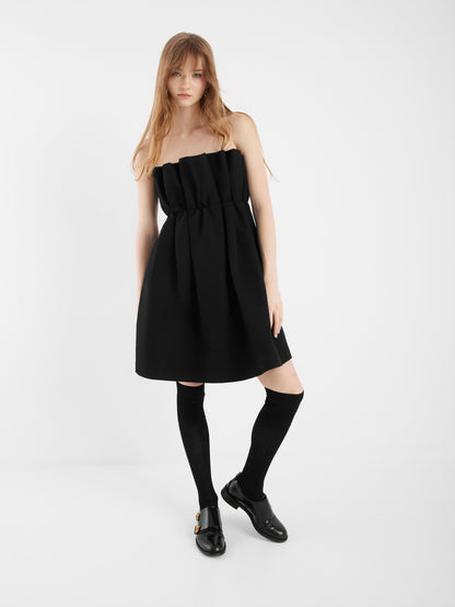 Eine Frau mit langen blonden Haaren trägt das Kleid Pleat black in einer A-Linien-Silhouette, gepaart mit kniehohen schwarzen Socken und schwarzen Schuhen, vor einem schlichten weißen Hintergrund.