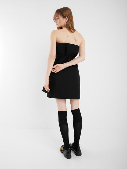 Eine Frau steht mit dem Gesicht zur Wand und trägt ein schwarzes Kleid mit eleganter A-Linien-Silhouette. Sie vervollständigt ihren Look mit schwarzen Kniestrümpfen und Schuhen und posiert mit hinter dem Rücken verschränkten Armen in einem minimalistischen Raum mit weißen Wänden.
