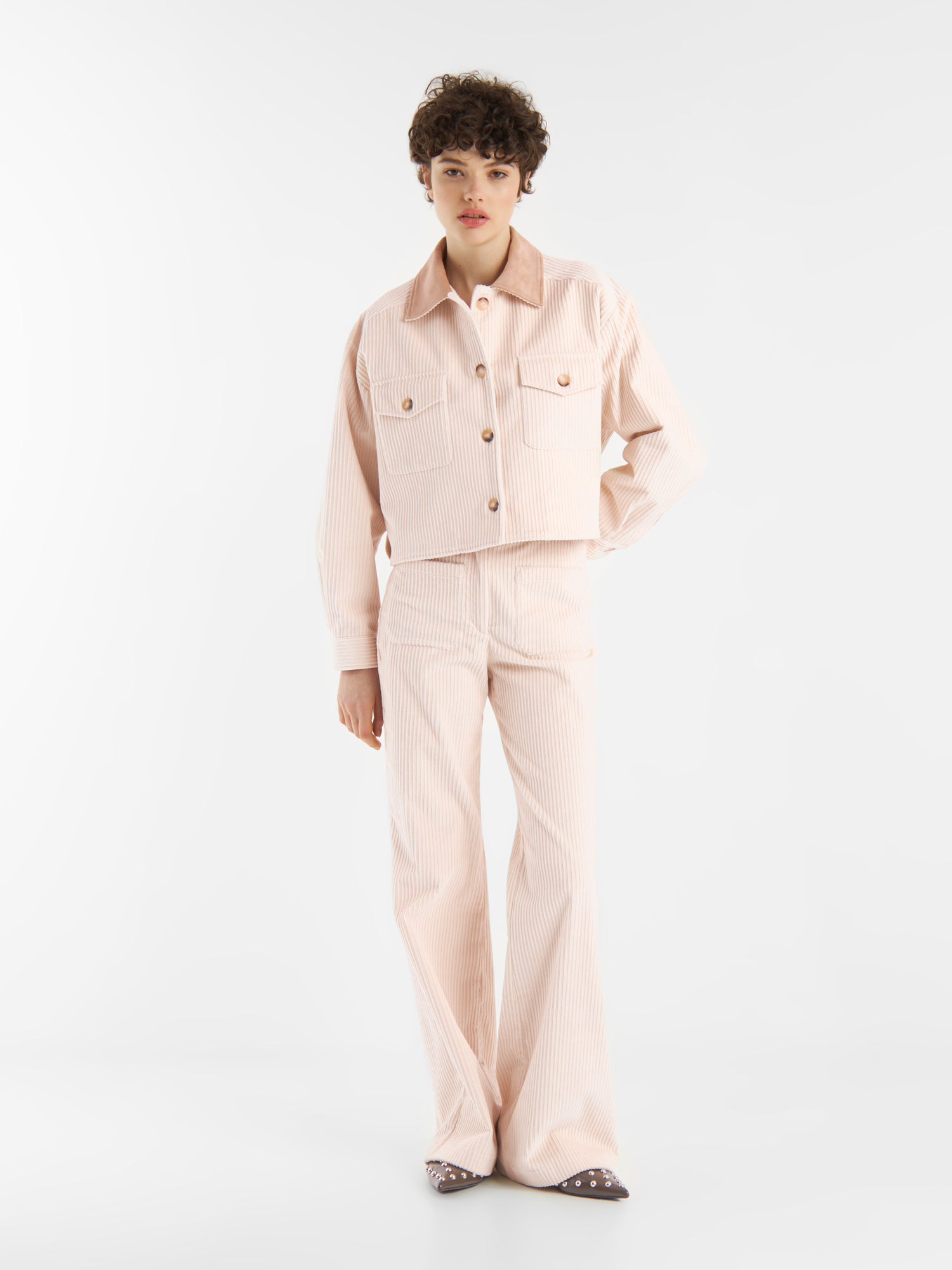 Eine Person mit kurzen lockigen Haaren trägt eine hellrosa, gestreifte Cordjacke OFW und eine dazu passende Schlaghose mit hoher Taille, die vor einem schlichten weißen Hintergrund steht.