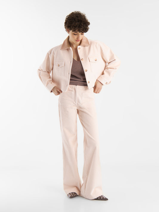 Eine Person mit kurzen lockigen Haaren trägt eine hellrosa Feincord-Jacke und eine dazu passende 70er-Jahre-Cord-Hose über einem lilafarbenen Oberteil und blickt mit den Händen in der Taille vor einem cremeweißen Hintergrund nach unten.