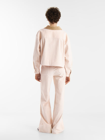 Person mit kurzen lockigen Haaren trägt eine hellrosa Cordjacke OFW und eine passende Schlaghose, steht vor einem schlichten weißen Hintergrund und blickt von der Kamera weg.