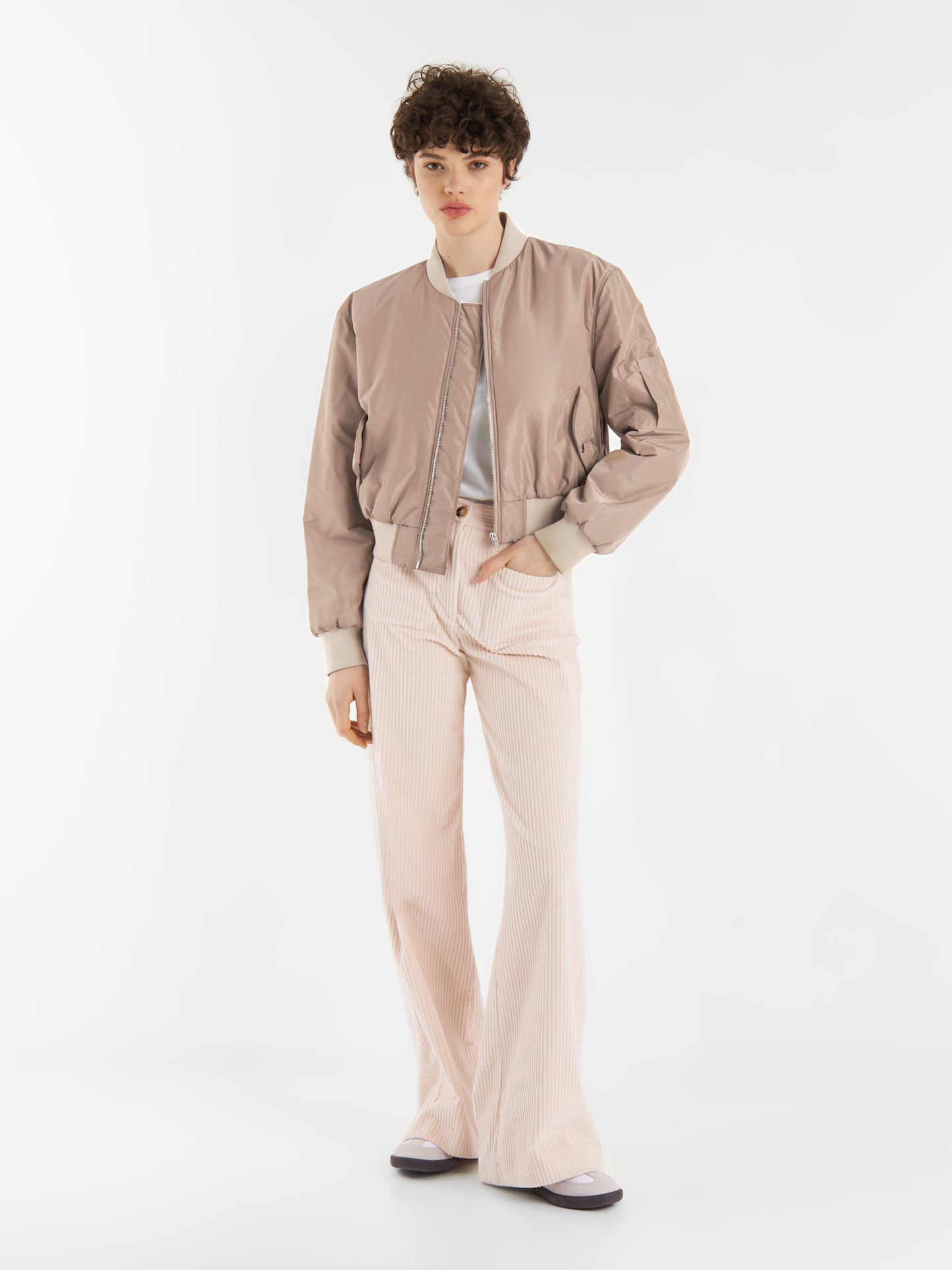 Eine Person mit kurzen lockigen Haaren trägt eine braune Bomberjacke, ein weißes Oberteil und eine hochtaillierte hellrosa Schlaghose, die vor einem schlichten weißen Hintergrund steht.