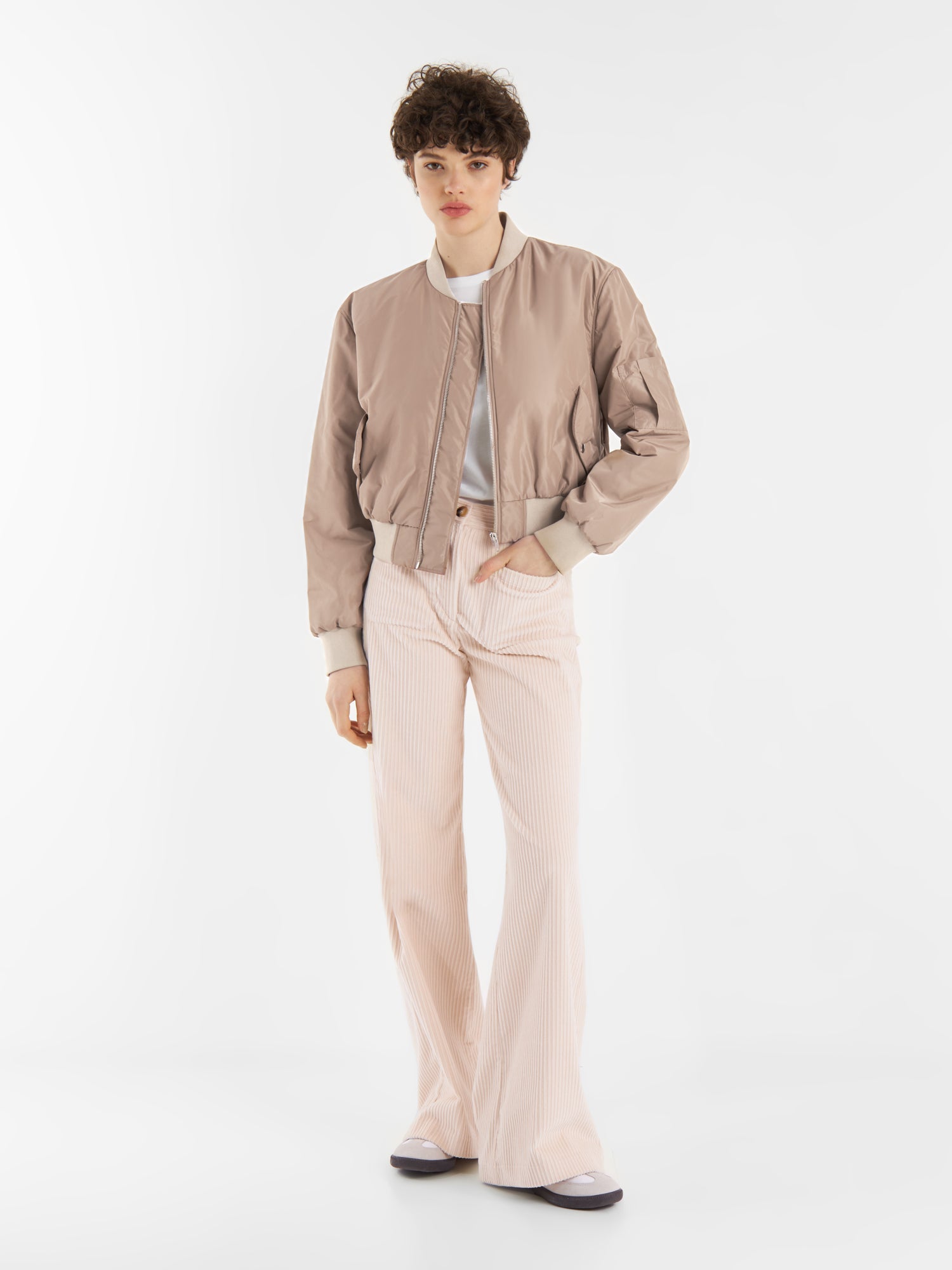 Eine Person mit kurzen lockigen Haaren trägt eine braune Bomberjacke, ein weißes Oberteil und eine hochtaillierte hellrosa Schlaghose, die vor einem schlichten weißen Hintergrund steht.