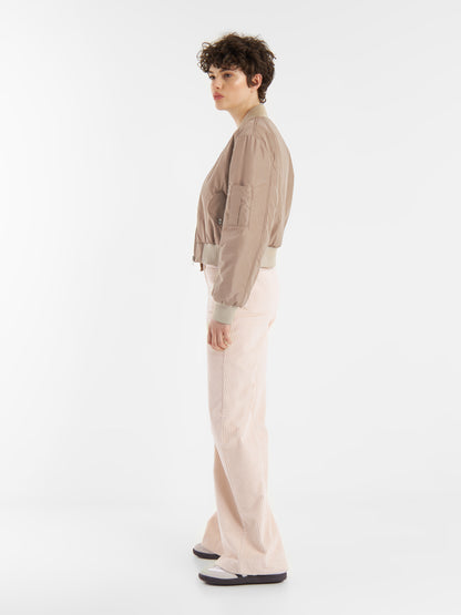 Eine Person mit kurzen lockigen Haaren steht seitlich vor einem schlichten weißen Hintergrund und trägt eine braune Bomberjacke, eine hellrosa Hose mit weitem Bein und schwarz-weiße Turnschuhe.