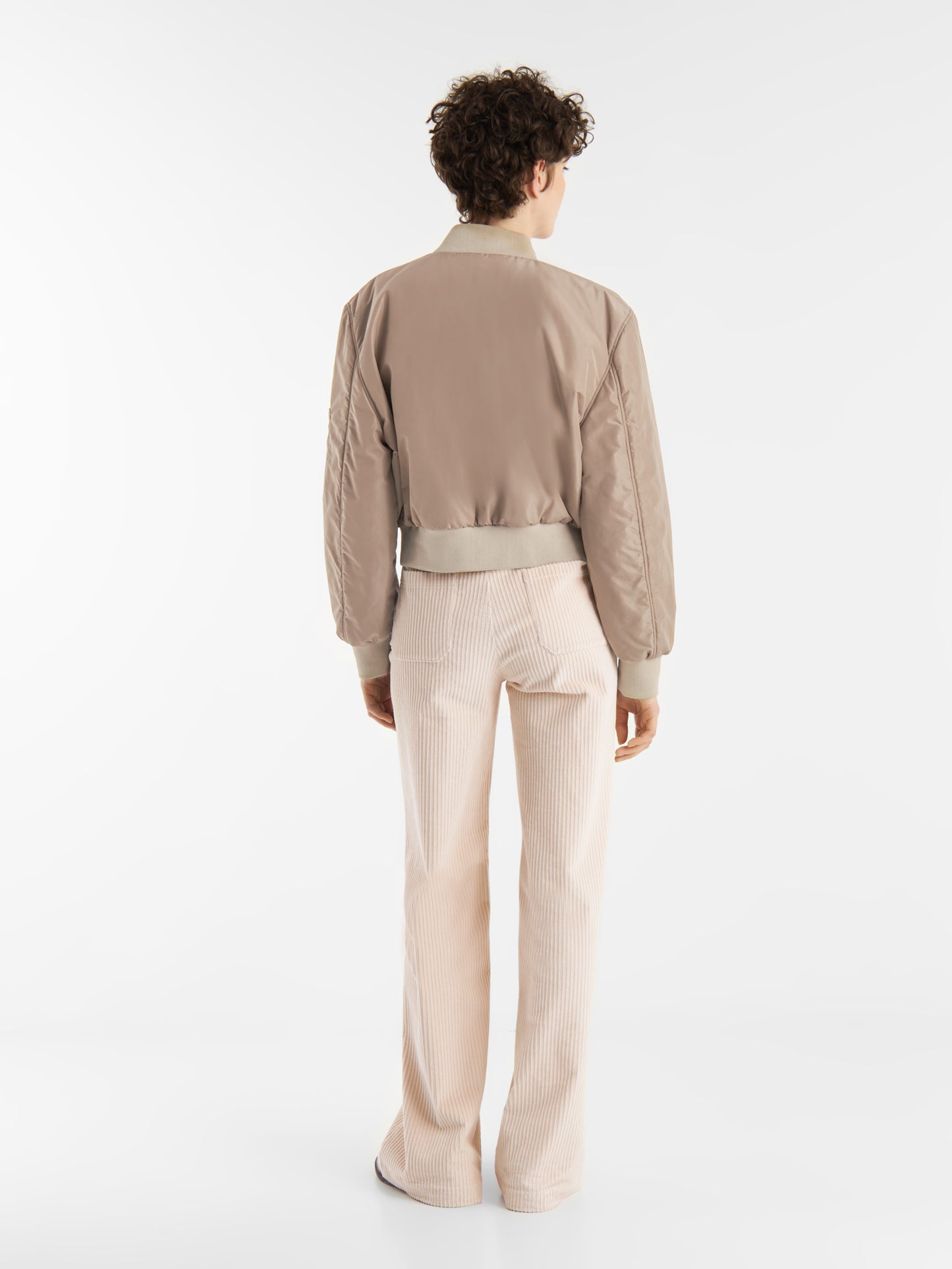 Eine Person mit kurzen lockigen Haaren steht mit dem Gesicht zur Wand und trägt die braune Jacke Bomber und eine cremefarbene Bundfaltenhose vor einem schlichten weißen Hintergrund.