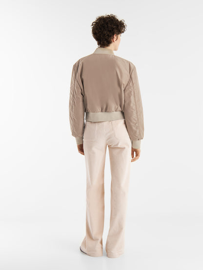 Eine Person mit kurzen lockigen Haaren steht mit dem Gesicht zur Wand und trägt die braune Jacke Bomber und eine cremefarbene Bundfaltenhose vor einem schlichten weißen Hintergrund.