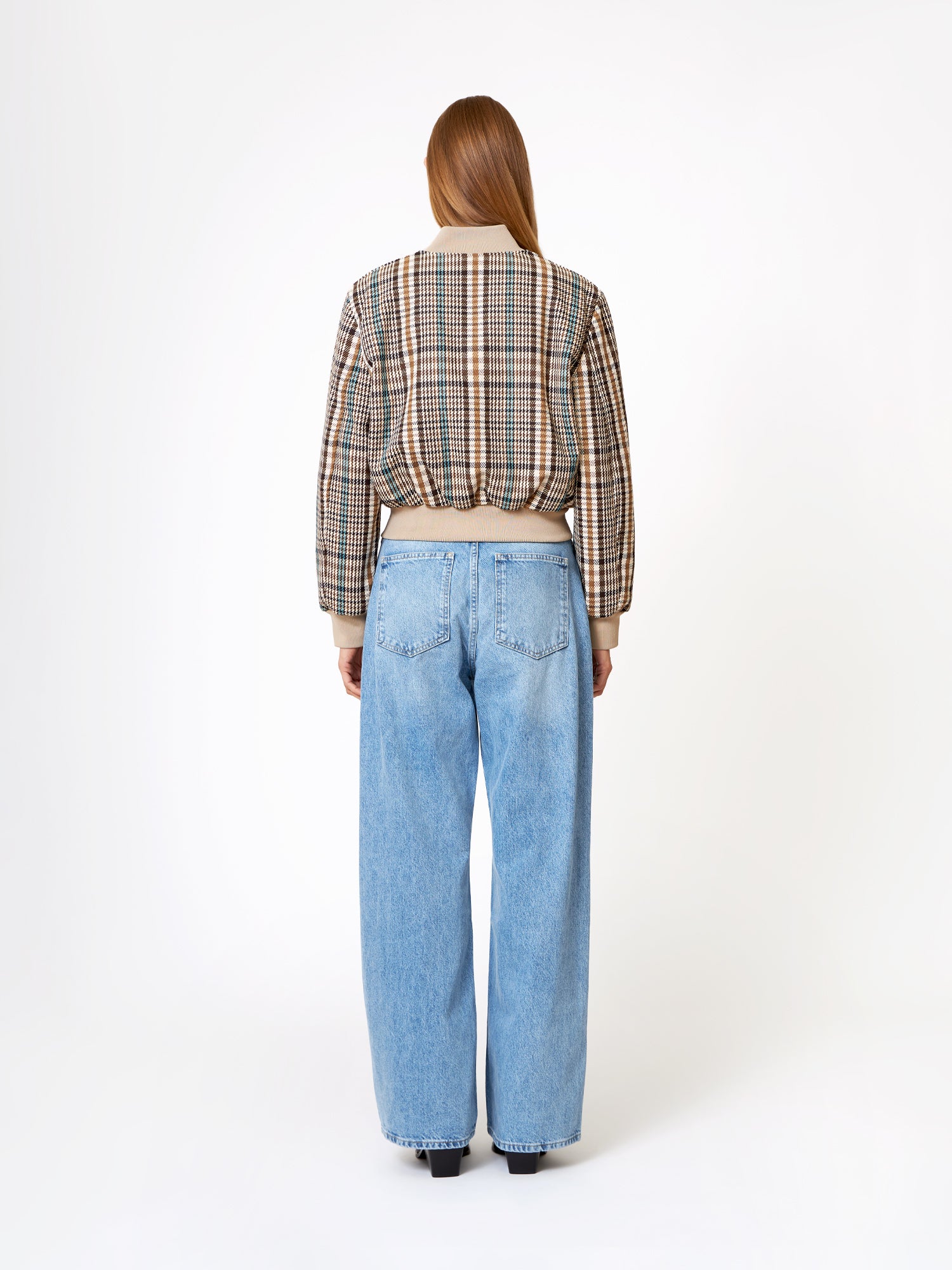 Eine Frau steht mit dem Gesicht zur Kamera, trägt eine Bomberjacke und eine hellblaue, hochgekrempelte Jeans mit weitem Bein vor einem schlichten weißen Hintergrund.