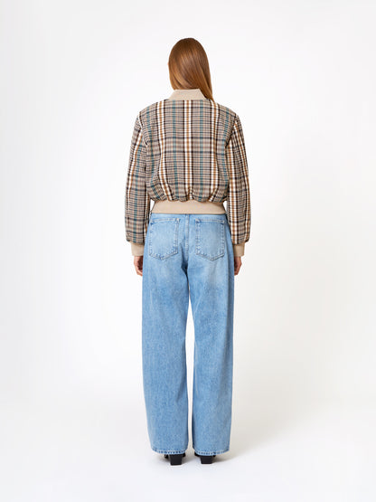 Eine Frau steht mit dem Gesicht zur Kamera, trägt eine Bomberjacke und eine hellblaue, hochgekrempelte Jeans mit weitem Bein vor einem schlichten weißen Hintergrund.