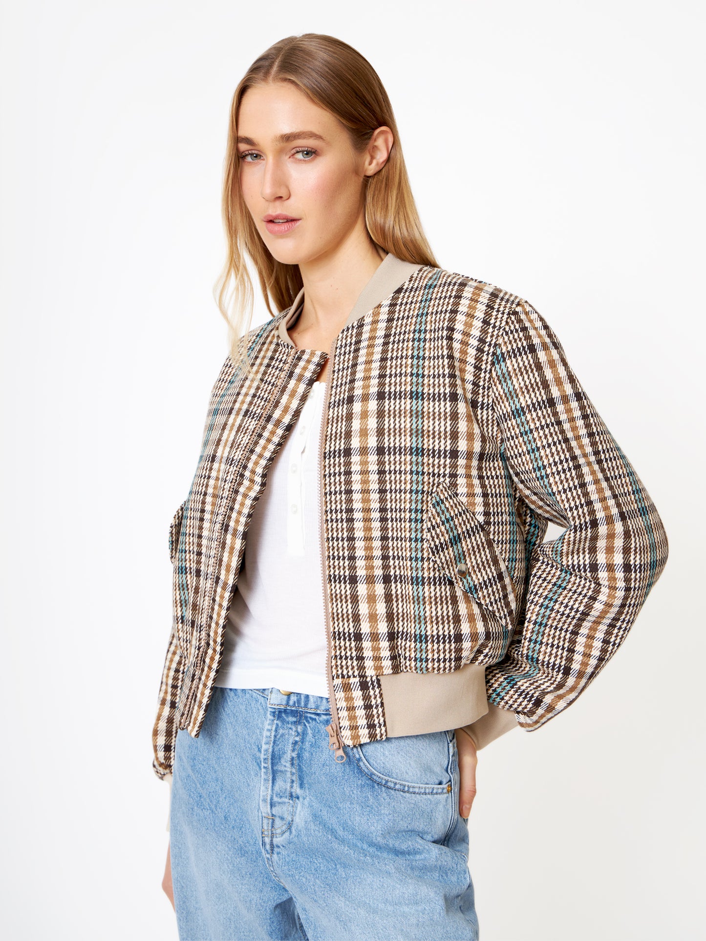 Eine Frau, die eine Bomberjacke aus braun kariertem Wollgemisch über einem weißen Hemd und hellblauen Jeans trägt, steht vor einem weißen Hintergrund, leicht zur Seite geneigt und mit einer Hand in der Tasche.
