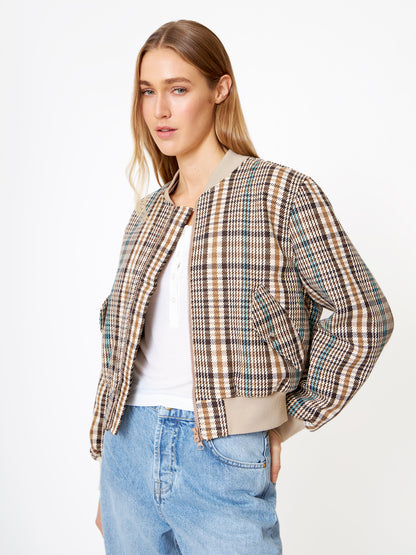 Eine Frau, die eine Bomberjacke aus braun kariertem Wollgemisch über einem weißen Hemd und hellblauen Jeans trägt, steht vor einem weißen Hintergrund, leicht zur Seite geneigt und mit einer Hand in der Tasche.
