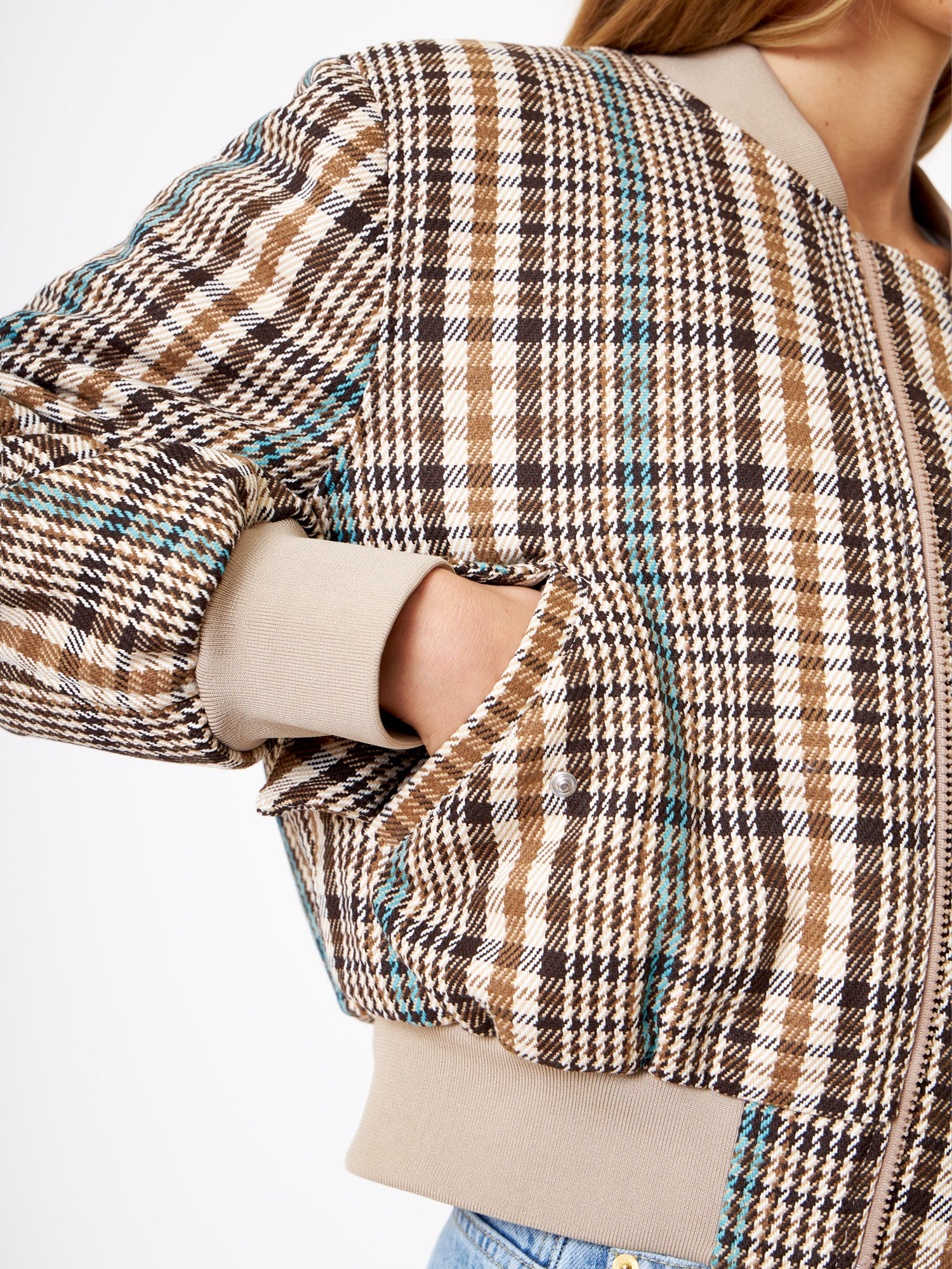 Eine Person in einer Bomberjacke mit braunem und beigem Karomuster und blaugrünen Akzenten steht mit einer Hand in der Tasche. Das Bild hebt das strukturierte Muster der Jacke hervor und konzentriert sich auf den Oberkörper und den Arm.