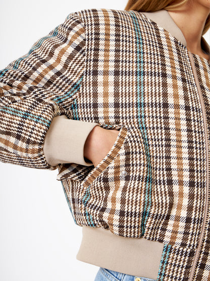 Eine Person in einer Bomberjacke mit braunem und beigem Karomuster und blaugrünen Akzenten steht mit einer Hand in der Tasche. Das Bild hebt das strukturierte Muster der Jacke hervor und konzentriert sich auf den Oberkörper und den Arm.