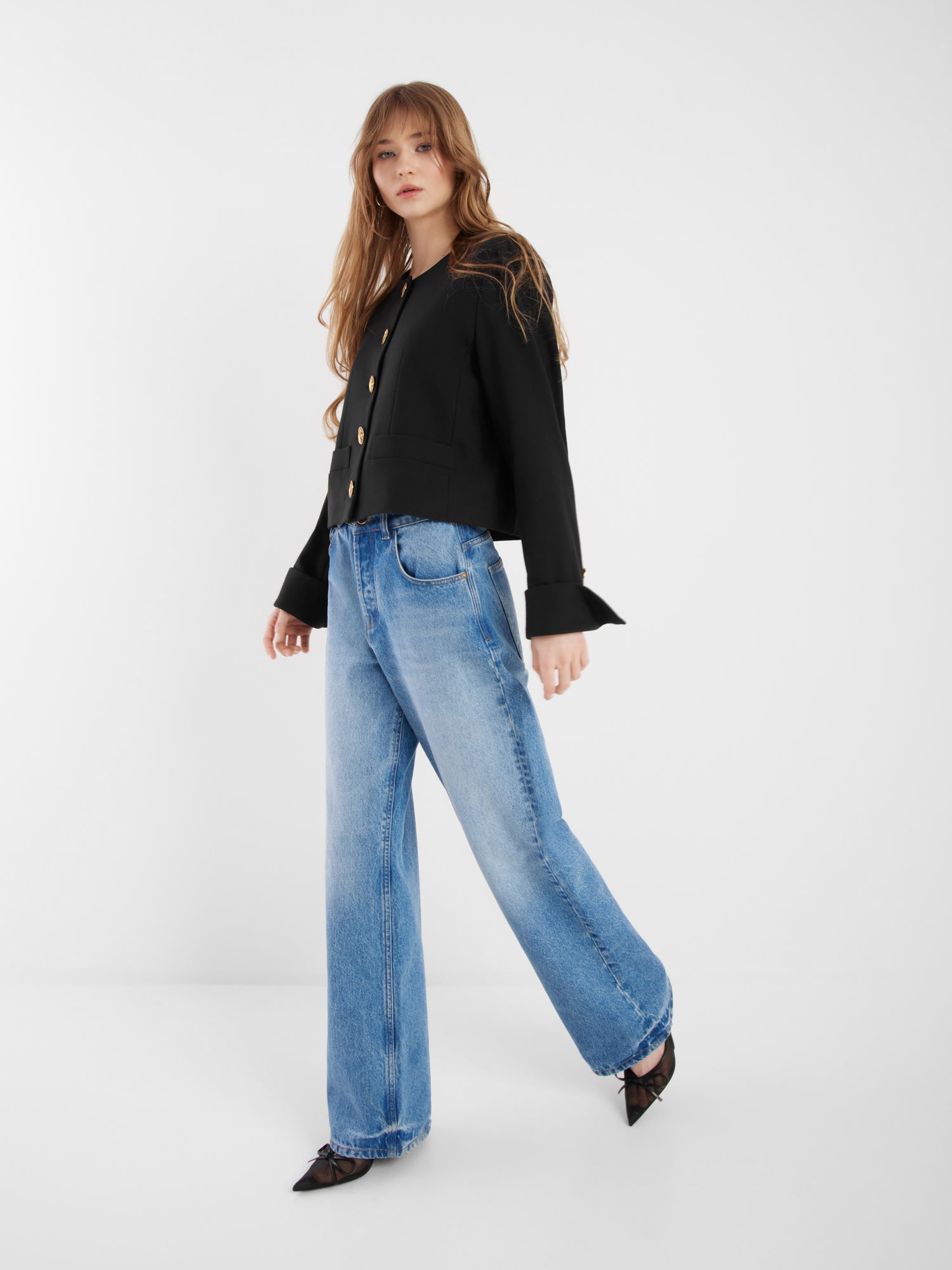 Eine Frau mit langen braunen Haaren trägt die schwarze Jacke Yves, hellblaue Jeans mit weitem Bein und spitze schwarze Schuhe und posiert vor einem schlichten weißen Hintergrund.