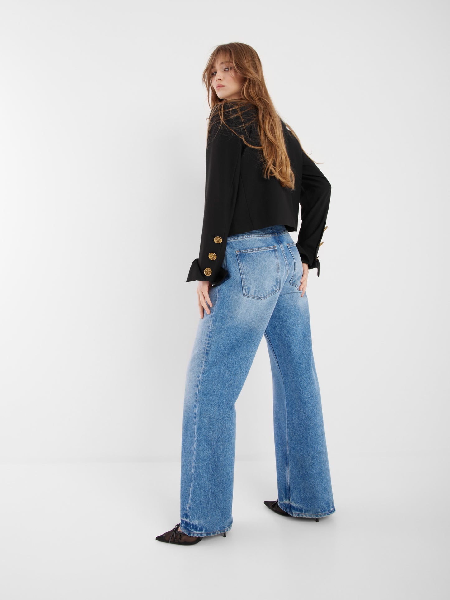 Eine Frau mit langen Haaren steht mit abgewandtem Gesicht und dreht den Kopf. Sie trägt die schwarze Jacke Yves, gepaart mit einer blauen Jeans mit hoher Taille und weitem Bein und schwarzen spitzen Schuhen in einem minimalen weißen Raum.