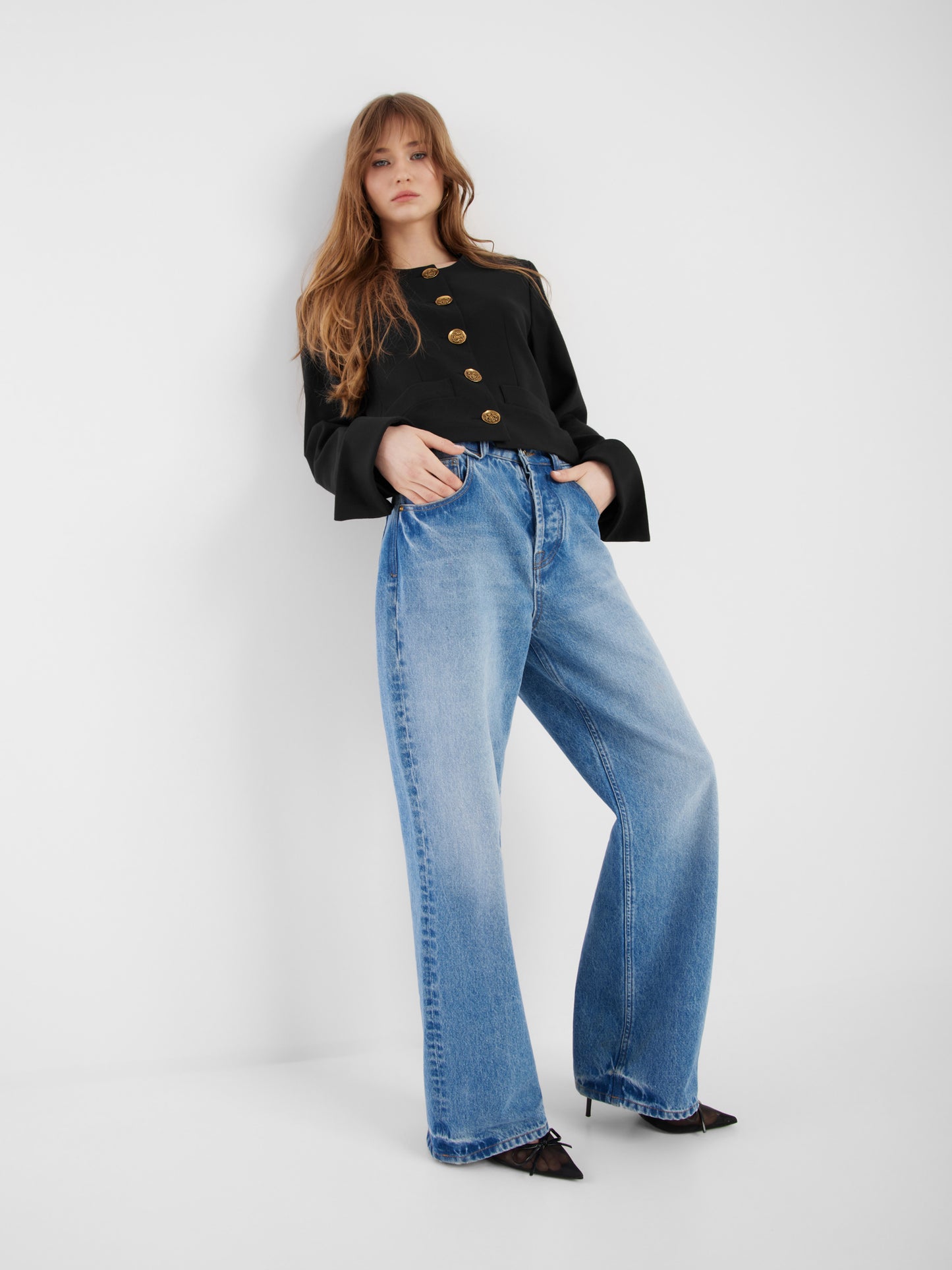 Eine Frau steht vor einer weißen Wand und trägt die schwarze Jacke Yves mit einer blauen Jeans mit hoher Taille und weitem Bein und schwarzen spitzen Absätzen. Ihre Hände stecken in den Taschen und ihr langes Haar ist offen.