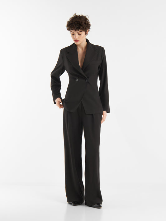 Eine Person mit kurzen lockigen Haaren modelliert den locker sitzenden schwarzen Anzug "Ready For Business", der aus einem doppelreihigen Blazer und einer Hose mit weitem Bein besteht, vor einem schlichten weißen Hintergrund.