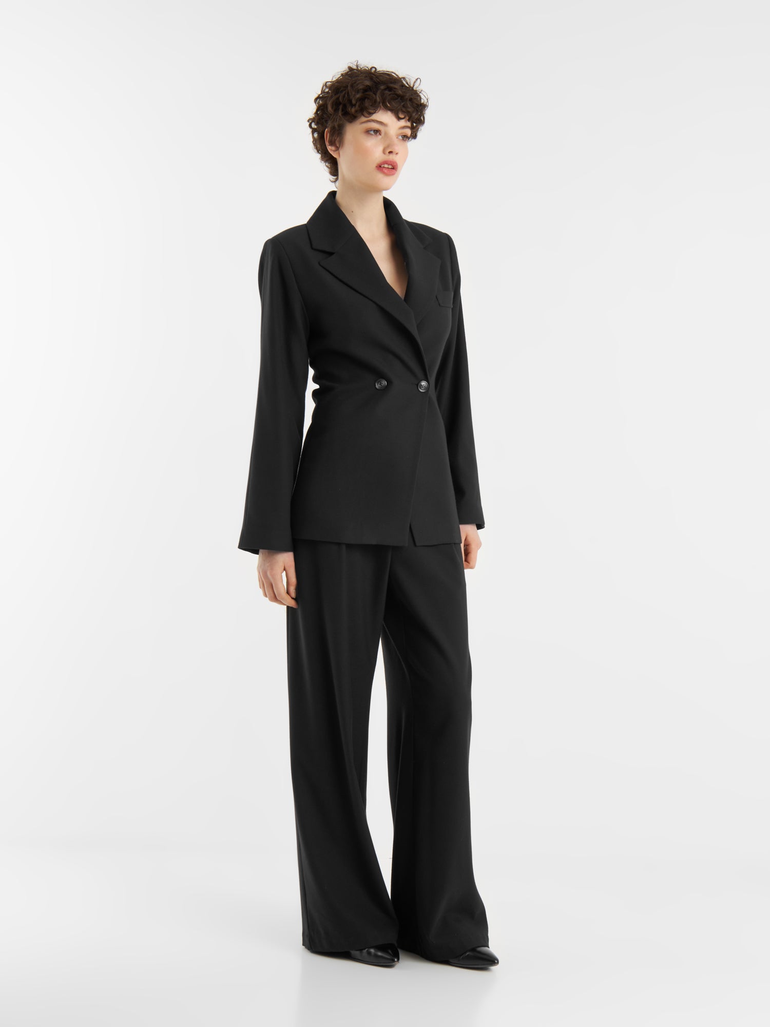Eine Person mit kurzen lockigen Haaren steht vor einem weißen Hintergrund und trägt das Set "Office & Evening": einen schwarzen Zweireiher, eine passende Hose mit weitem Bein und schwarze Schuhe.