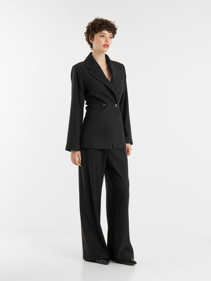 Eine Person mit kurzen lockigen Haaren steht vor einem weißen Hintergrund und trägt das Set "Office & Evening": einen schwarzen Zweireiher, eine passende Hose mit weitem Bein und schwarze Schuhe.