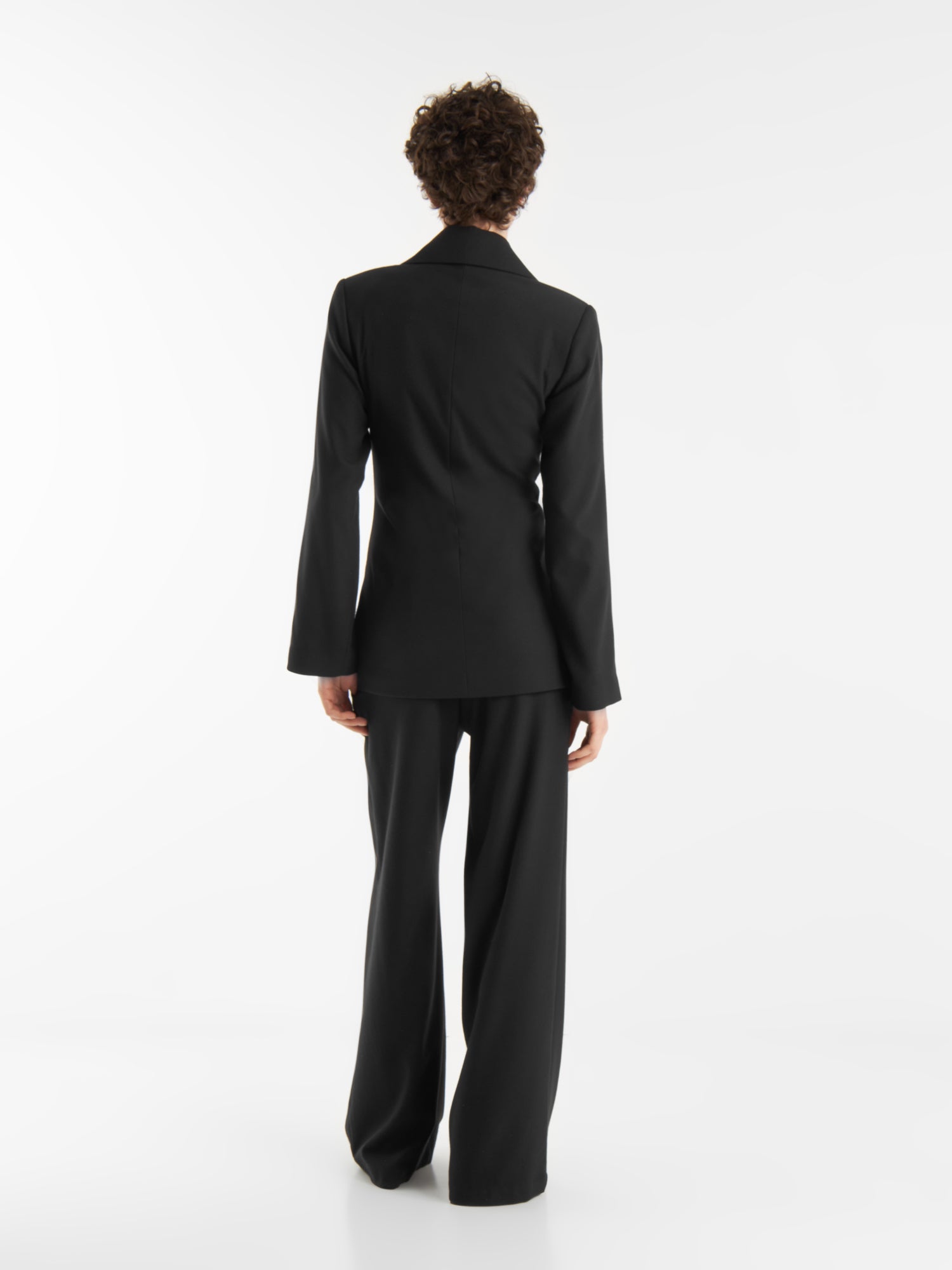 Eine Person mit kurzen lockigen Haaren, die die schwarze Anzugsjacke und -hose "Office & Evening" trägt, steht mit dem Gesicht nach außen vor einem schlichten weißen Hintergrund.