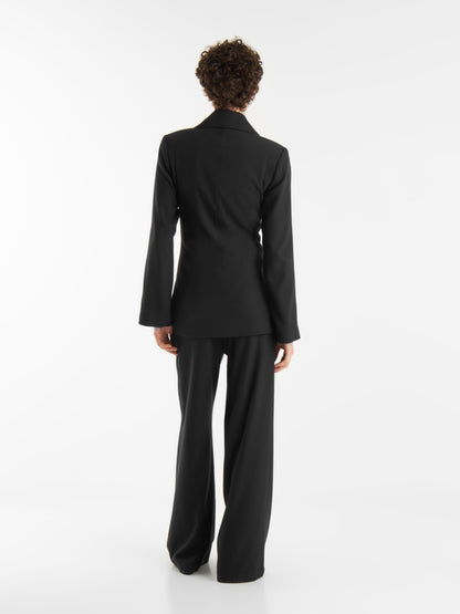 Eine Person mit kurzen lockigen Haaren, die die schwarze Anzugsjacke und -hose "Office & Evening" trägt, steht mit dem Gesicht nach außen vor einem schlichten weißen Hintergrund.