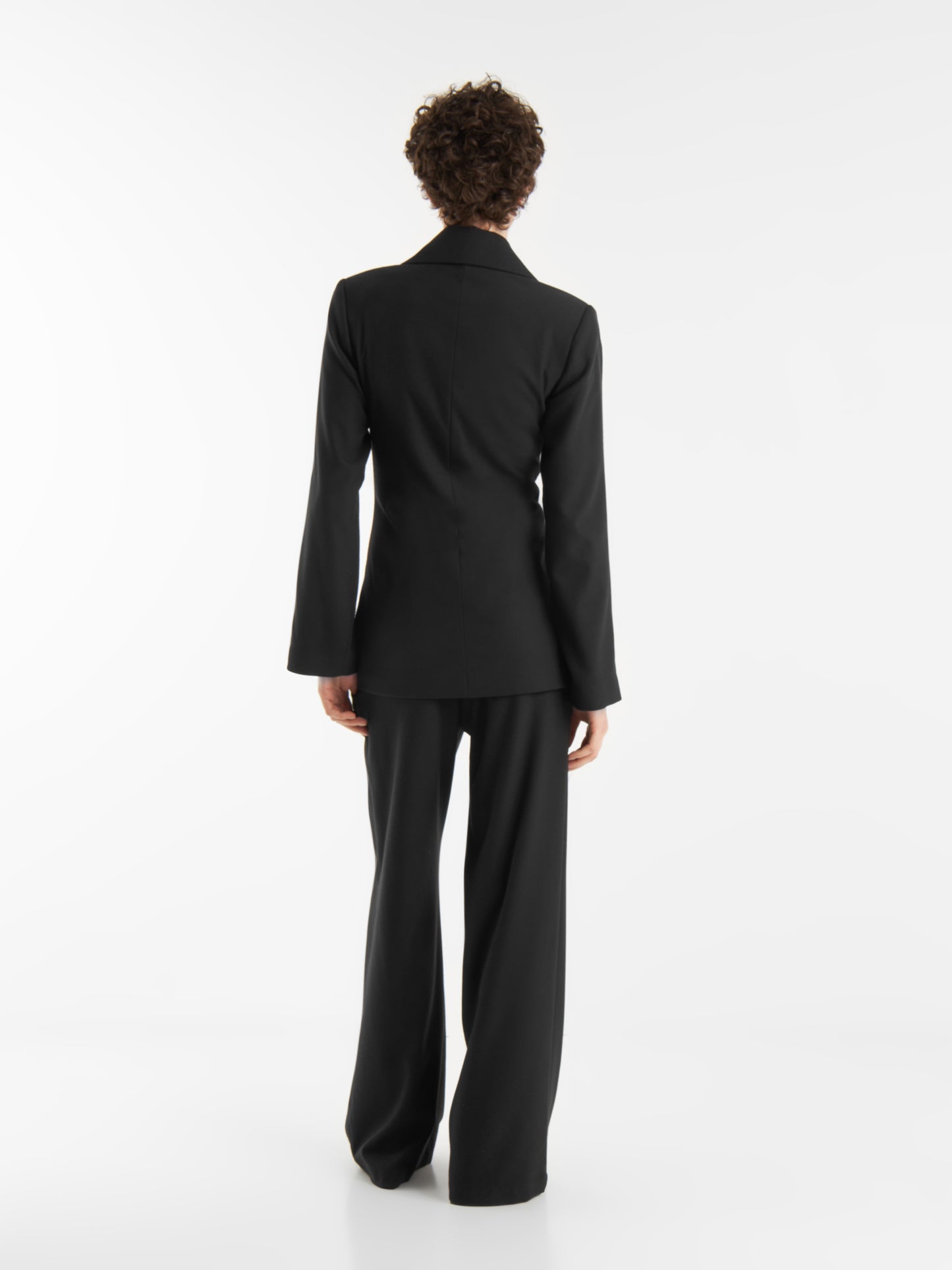 Eine Person mit kurzen lockigen Haaren steht mit dem Gesicht zur Wand, trägt einen schwarzen maßgeschneiderten Blazer und die Hose Boyfriend schwarz, vor einem schlichten weißen Hintergrund.
