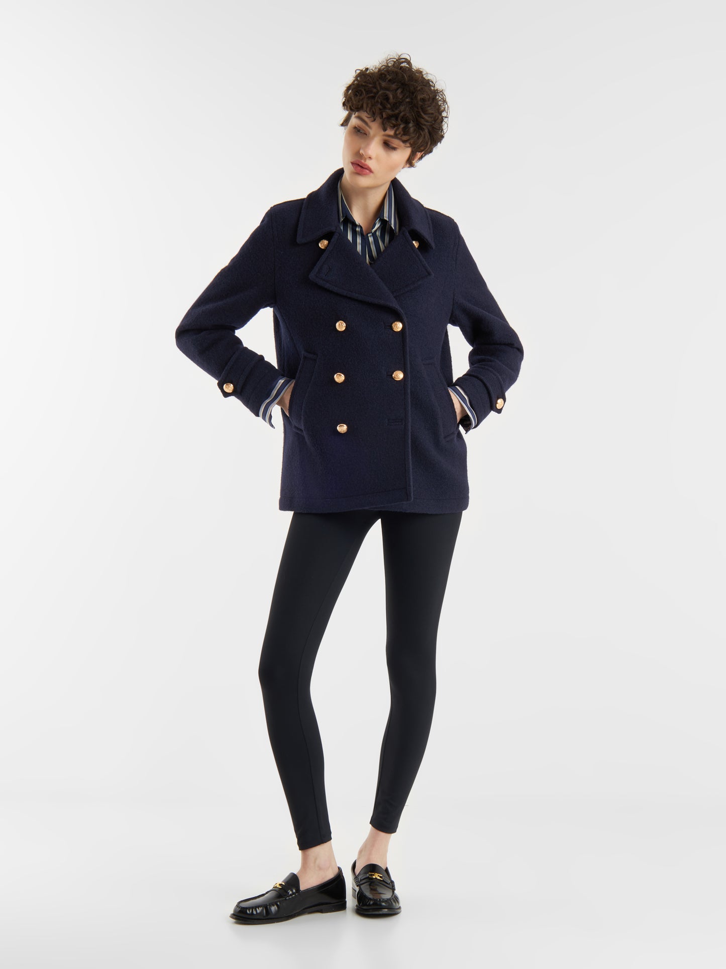 Eine Person mit kurzen lockigen Haaren trägt die Jacke Caban Navy Gold im klassischen Marine-Look, gepaart mit einem gestreiften Hemd, schwarzen Leggings und schwarzen Halbschuhen, vor einem schlichten weißen Hintergrund.