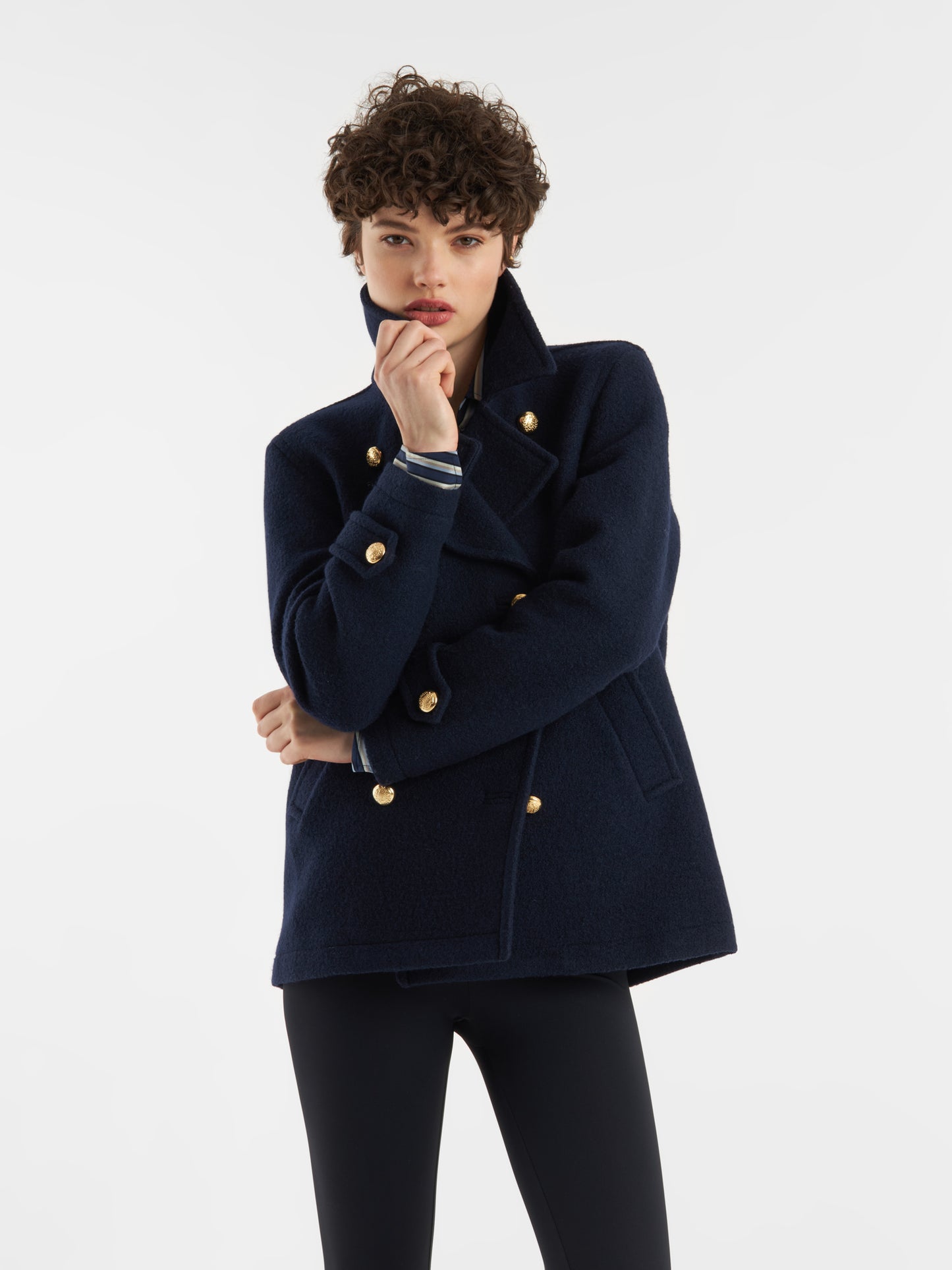 Eine Person mit kurzen lockigen Haaren trägt die Jacke Caban Navy Gold mit einer dunklen Hose vor einem schlichten weißen Hintergrund, eine Hand am Kinn und den anderen Arm gekreuzt für einen zeitlosen, von der Marine inspirierten Look.