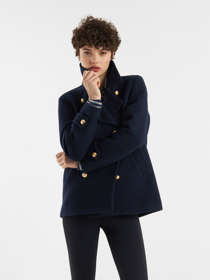 Eine Person mit kurzen lockigen Haaren trägt die Jacke Caban Navy Gold mit einer dunklen Hose vor einem schlichten weißen Hintergrund, eine Hand am Kinn und den anderen Arm gekreuzt für einen zeitlosen, von der Marine inspirierten Look.