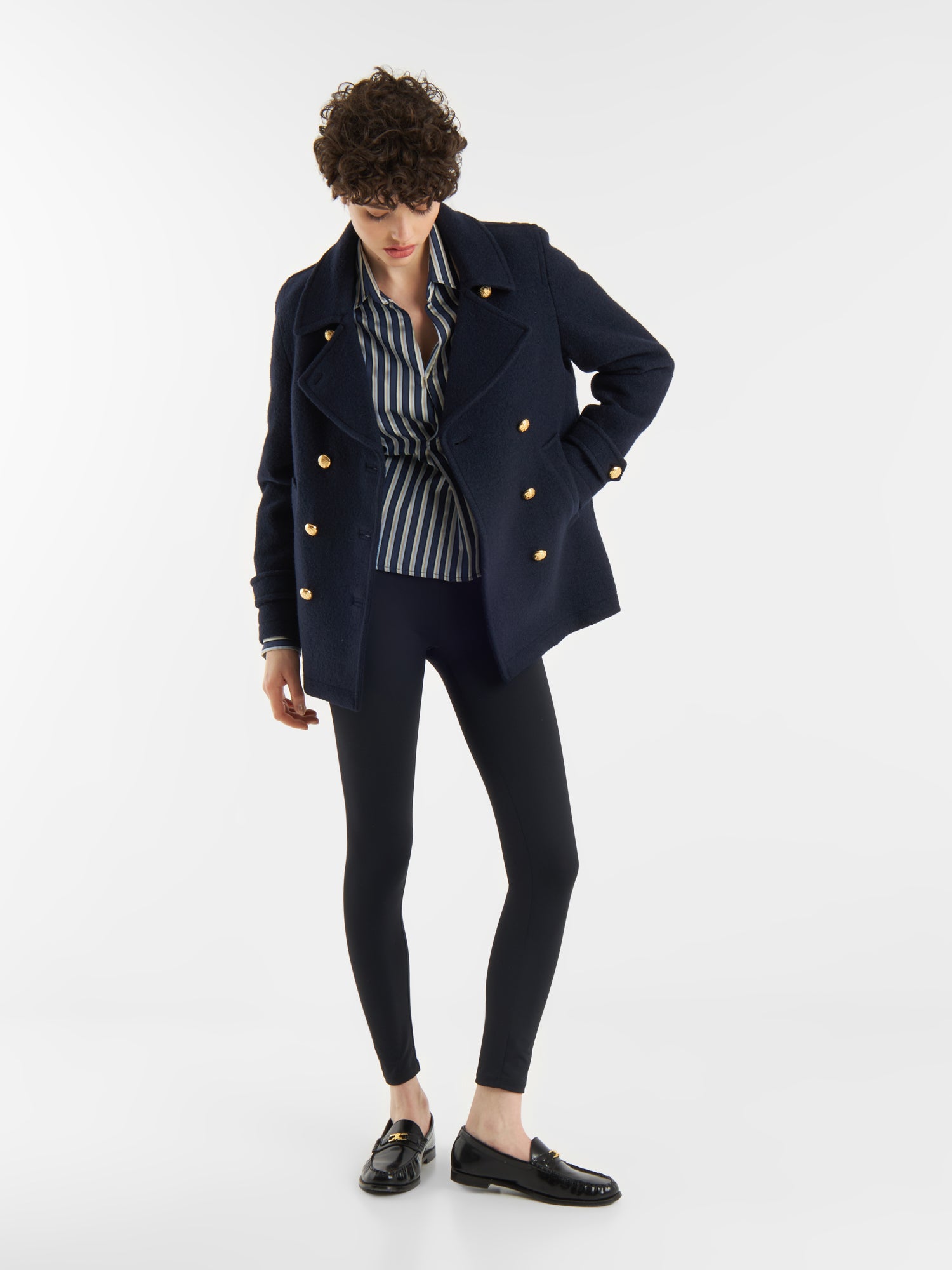 Eine Person mit kurzen lockigen Haaren modelliert die Jacke Caban Navy Gold mit einem gestreiften Hemd, schwarzen Leggings und Halbschuhen vor einem weißen Hintergrund für einen klassischen Marine-Look.