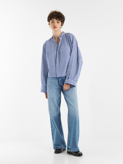 Person mit kurzen lockigen Haaren, die den Bluse Bomber Stripe Blue white (Oversize-Hemd aus 100% Baumwolle), hellblaue Jeans und schwarze Schuhe trägt, stehend vor einem schlichten weißen Hintergrund.