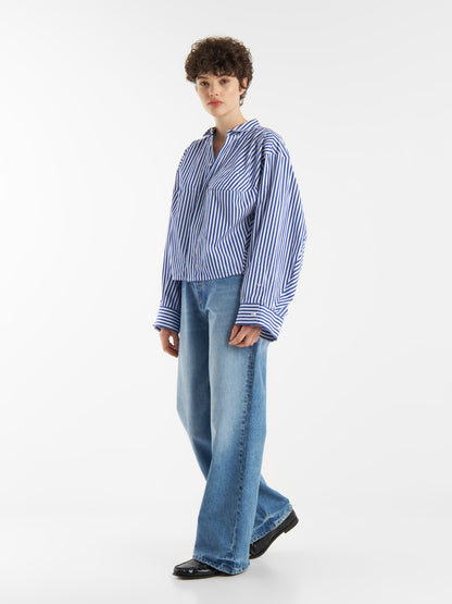 Eine Person mit kurzen lockigen Haaren trägt den Bluse Bomber Stripe Blue White und weite Blue Jeans, stehend und nach vorne blickend vor einem weißen Hintergrund.