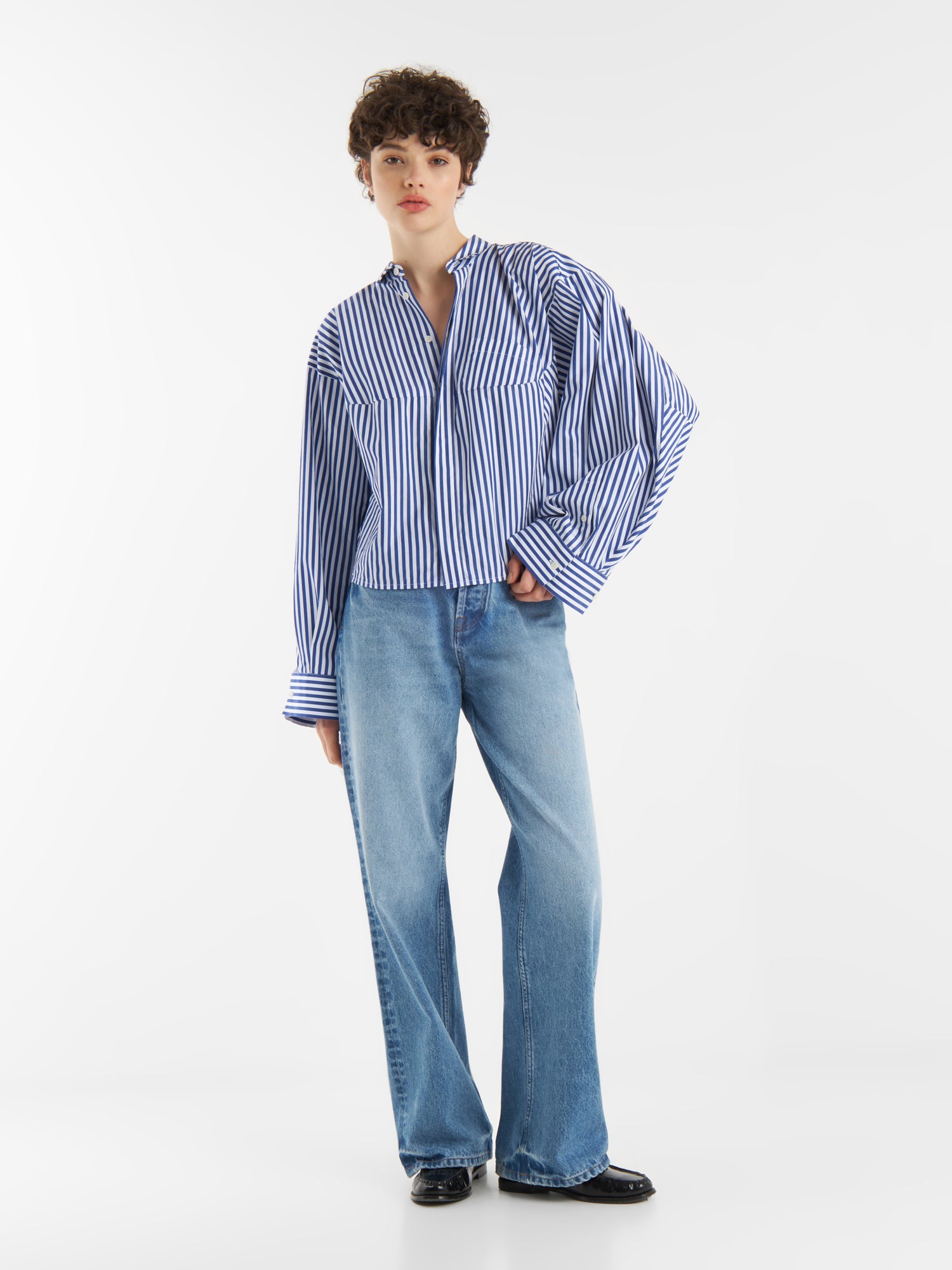 Eine Person mit kurzen lockigen Haaren trägt die Bluse Bomber Blue White Stripes, gepaart mit blauen Jeans und schwarzen Schuhen, vor einem schlichten weißen Hintergrund.