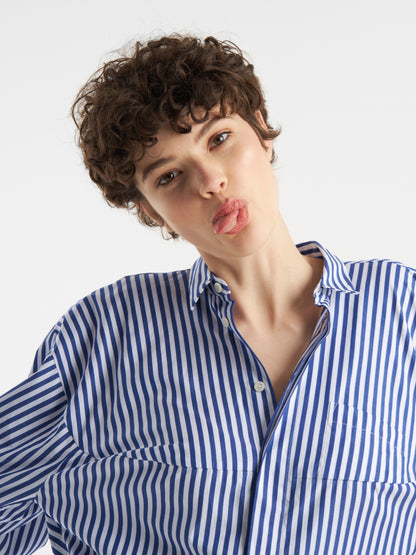 Eine Person mit kurzen lockigen Haaren, die den Bluse Bomber Stripe Blue white trägt, streckt spielerisch ihre Zunge vor einem schlichten weißen Hintergrund heraus.