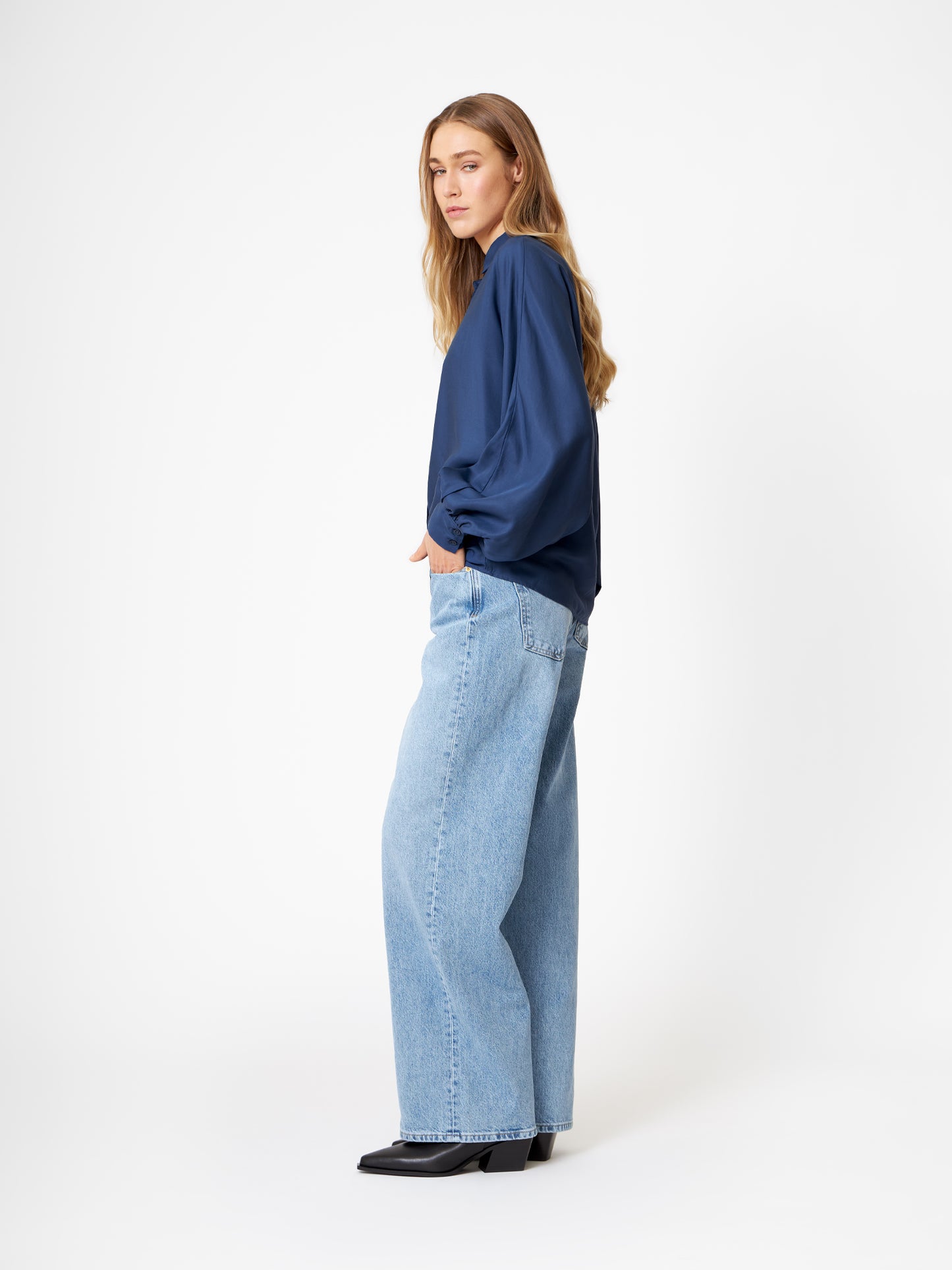 Eine Frau mit langen blonden Haaren steht seitlich vor einem weißen Hintergrund und trägt die Bluse Antonia Slim Tencel Blue, hellblaue Jeans mit weitem Bein und schwarze Schuhe.